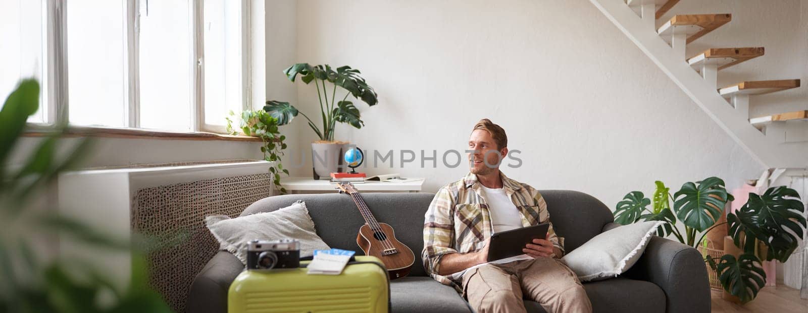 Image of young man with tablet, sitting in living room with suitcase, reading, studying the route, preparing for a vacation. Tourism concept