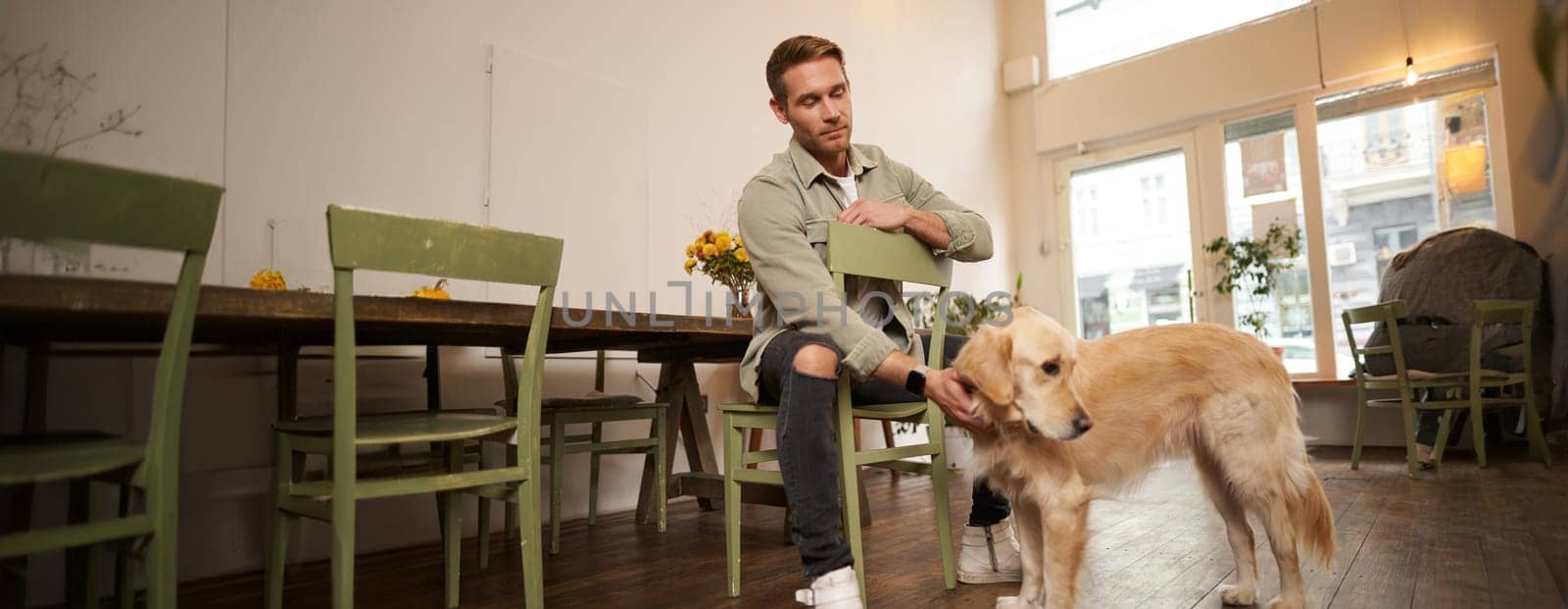 Close up portrait of handsome young man waiting for his order in coffee shop, petting a dog, resting in pet-friendly cafe.