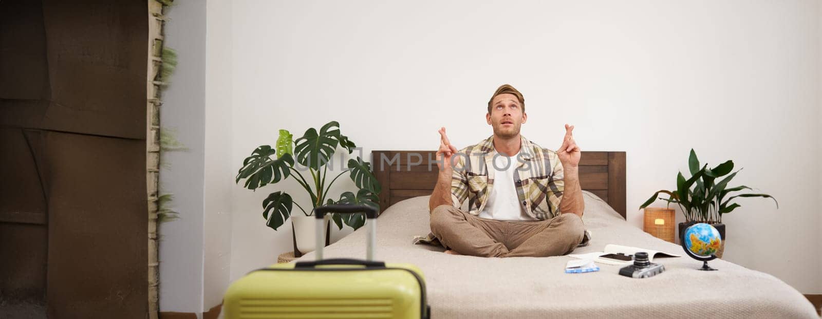 Portrait of young man meditating with fingers crossed, praying, making a wish, packing suitcase to go on holiday, has globe and camera on a bed.