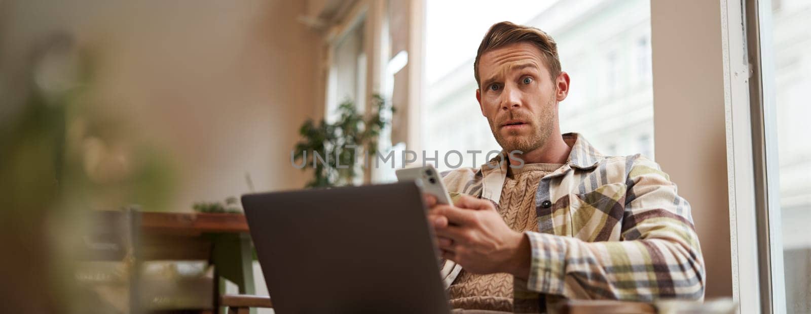 Portrait of cafe visitor sitting with laptop and mobile phone, looking confused, puzzled at camera. Lifestyle and public space concept