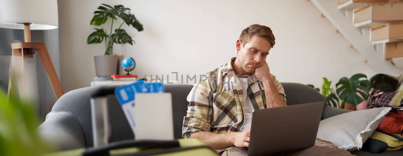 Portrait of gloomy, sad young man, has laptop opened, two plane tickets abroad and passport, looks disappointed at screen. Tourism concept