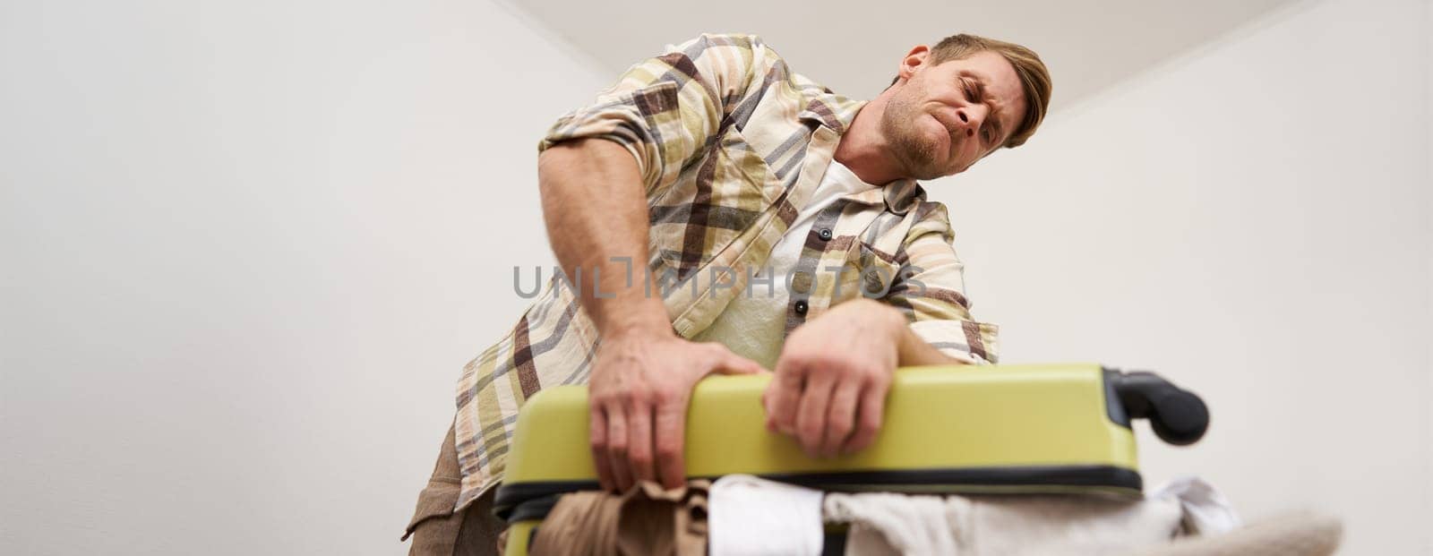 Image of young man, tourist struggling to close his suitcase full of clothes, pushing on bag, trying to press items inside luggage to shut the zipper, packing for travelling abroad, going on holiday,
