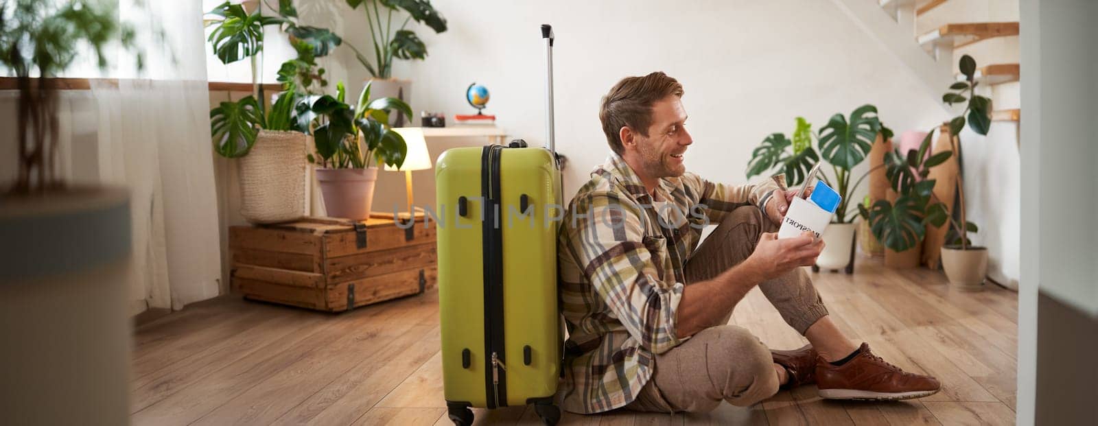 Portrait of smiling handsome man, sitting with suitcase on floor, holding passport and flight tickets, going on tour vacation.