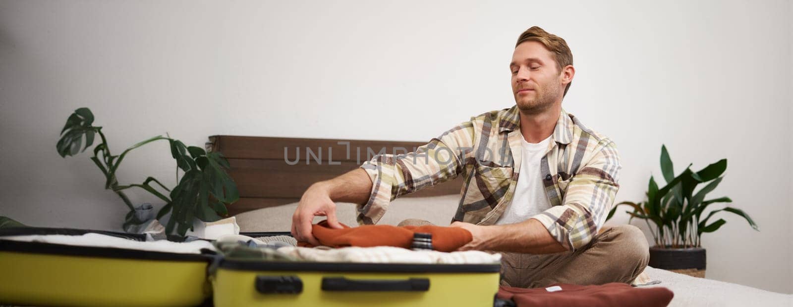 Portrait of handsome young man, tourist unpacking clothes in hotel room, staying in hostel on vacation, travelling, sitting with luggage on bed.