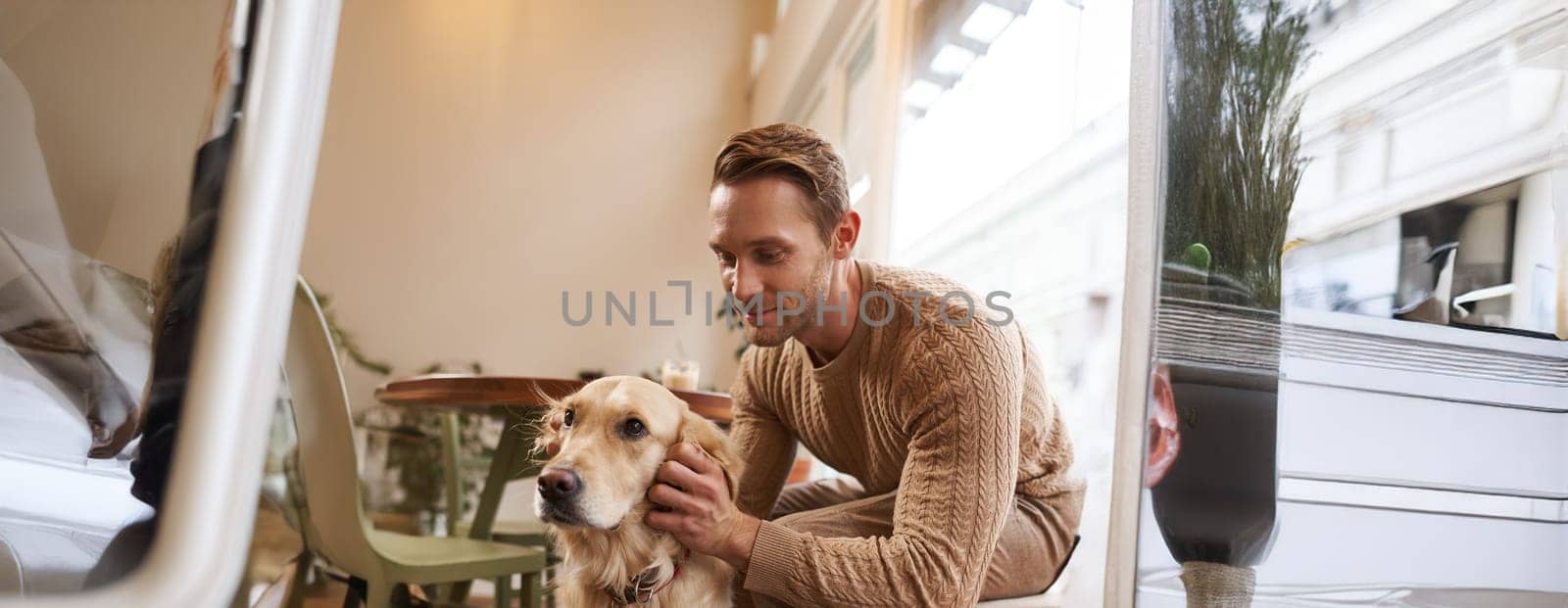 Close up portrait of handsome man with his dog, sitting in cafe. A guy drinks his coffee and touches golden retriever.