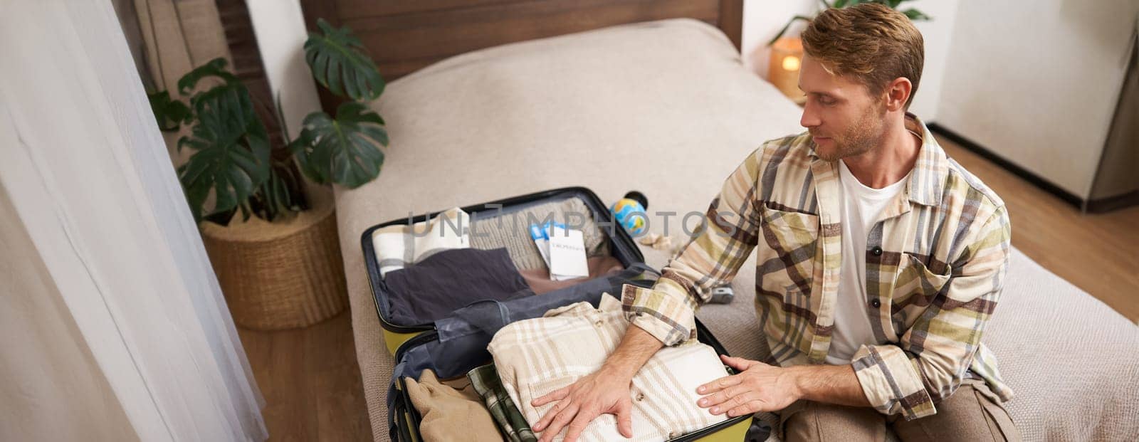 Portrait of young handsome man, tourist going on vacation, packing luggage for holidays, putting clothes in suitcase, sitting on bed.
