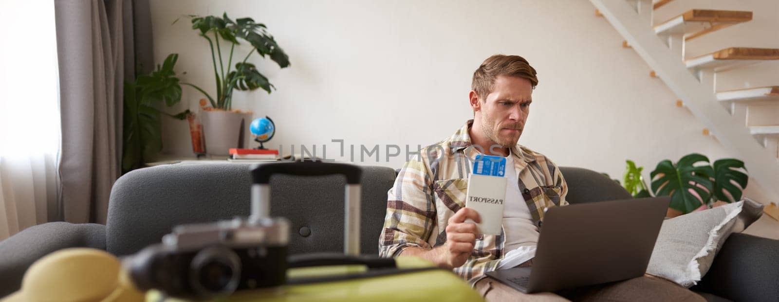 Angry man holding his flight tickets, staring upset at laptop screen, having problems with his holiday tour.