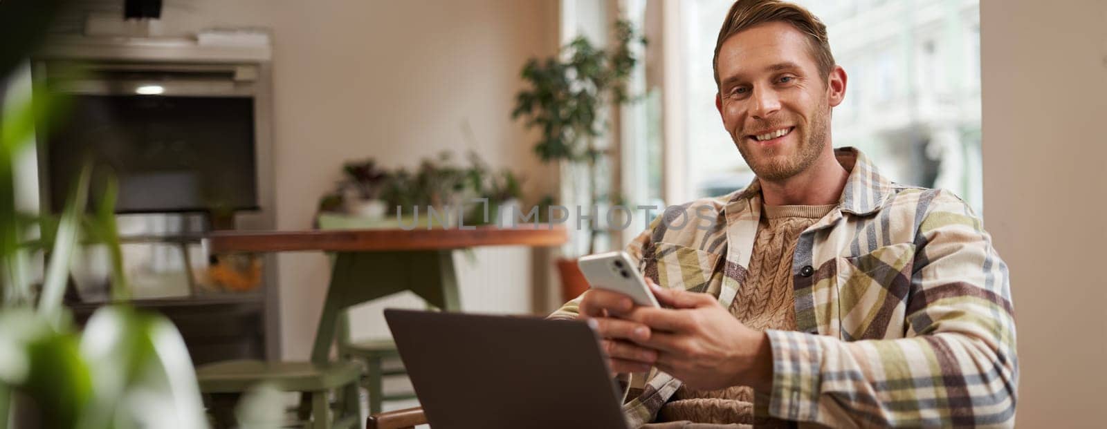 Portrait of young good-looking man, cafe visitor, sitting with laptop and smartphone, smiling at camera, freelancer working remotely from co-working space.