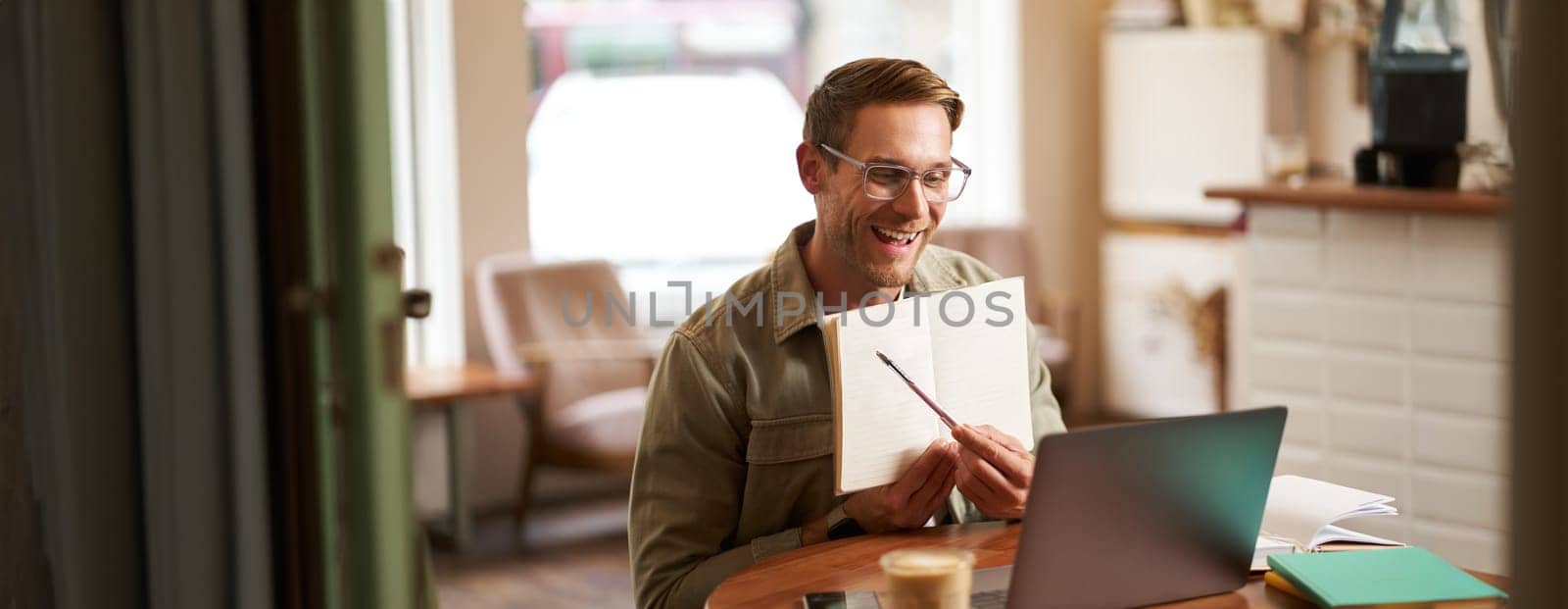 Portrait of young handsome man in glasses, private tutor teaching student online, pointing at his notebook, showing something, video chats via laptop application, working remotely in cafe.