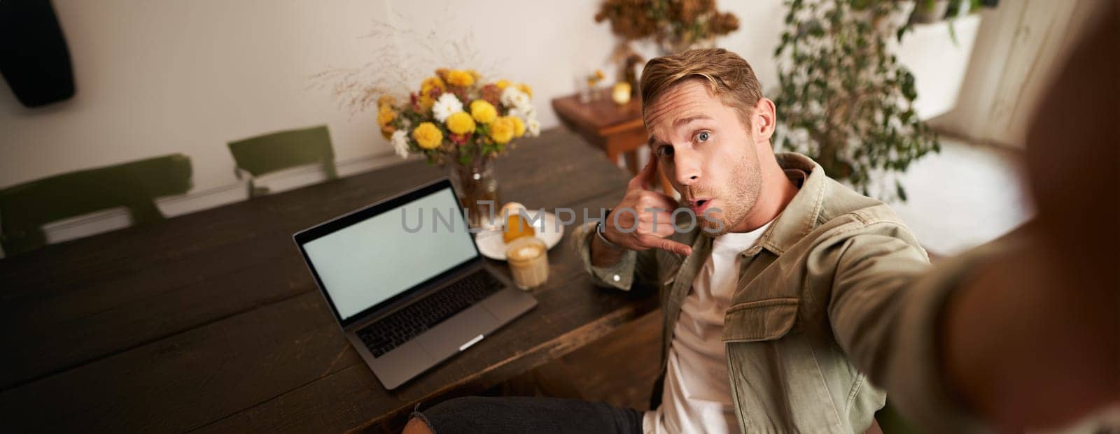 Handsome stylish young man takes selfie in cafe while working or studying remotely with laptop, shows call phone hand sign and smiling at mobile phone camera.