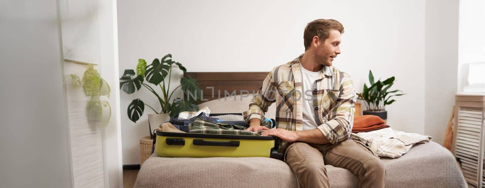 Tourism and travelling concept. Portrait of handsome young man going on holiday, packing his suitcase, checking luggage, sitting on bed and looking at packed things.