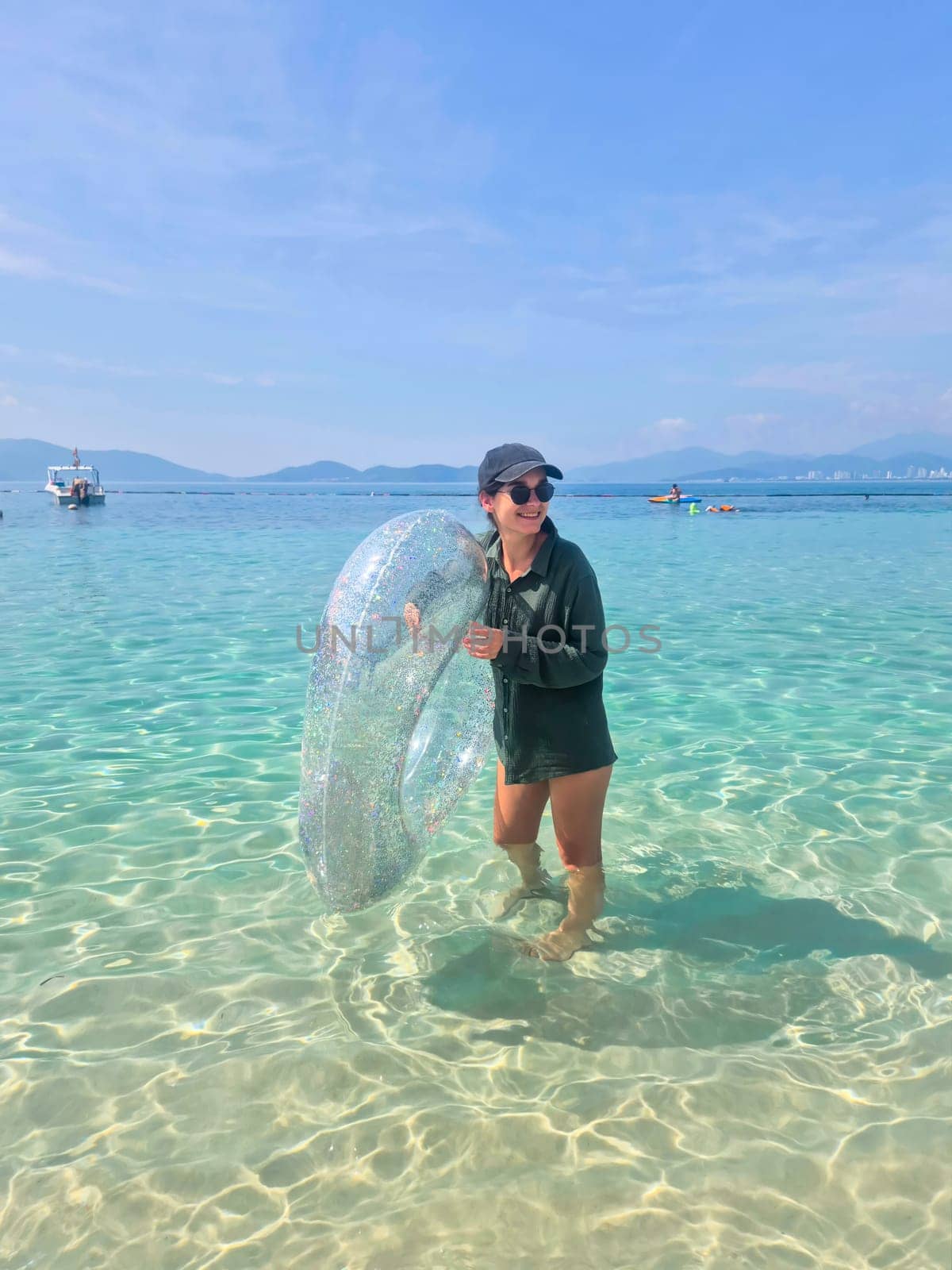 Brunette Russian girl in black swimsuit, sunglasses and shirt holds glittery pool float while laughing in Nha Trang waters. Sunny Vietnam beach day.