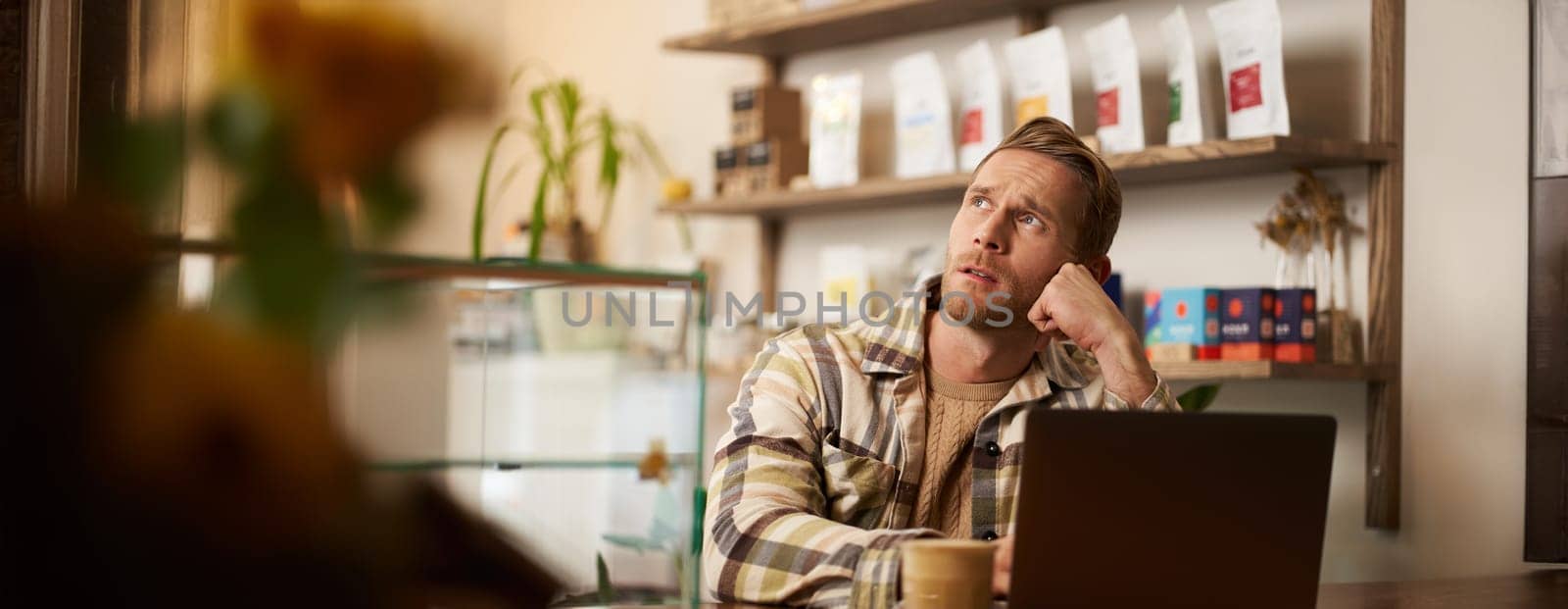 Man looking concentrated while working remotely from a coffee shop. Business owner sitting in cafe with laptop, drinking cappuccino, looking aside with thoughtful face, getting inspiration.
