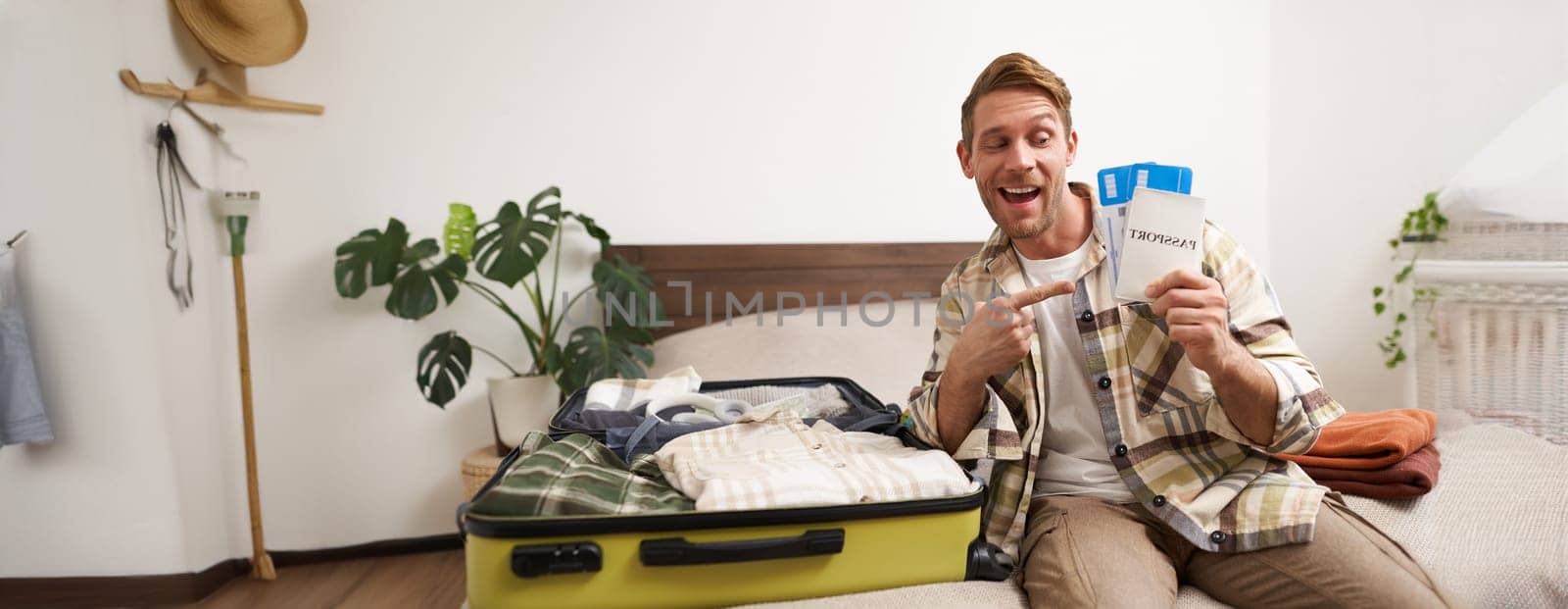 Portrait of handsome young man, traveller shows his passport and two tickets, packing suitcase with clothes, going on vacation. Tourism and holiday concept
