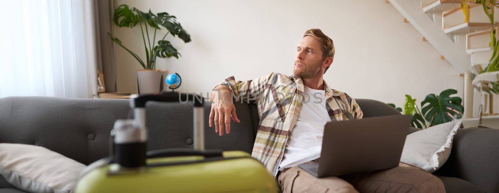 Happy man booking holiday online on laptop, paying for vacation on travel agency website, buying flight tickets in internet, sitting in living room with suitcase.