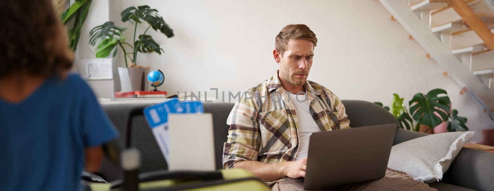 Portrait of serious and concentrated man, sits with laptop in living room, has two plane tickets, plans holiday, goes online.