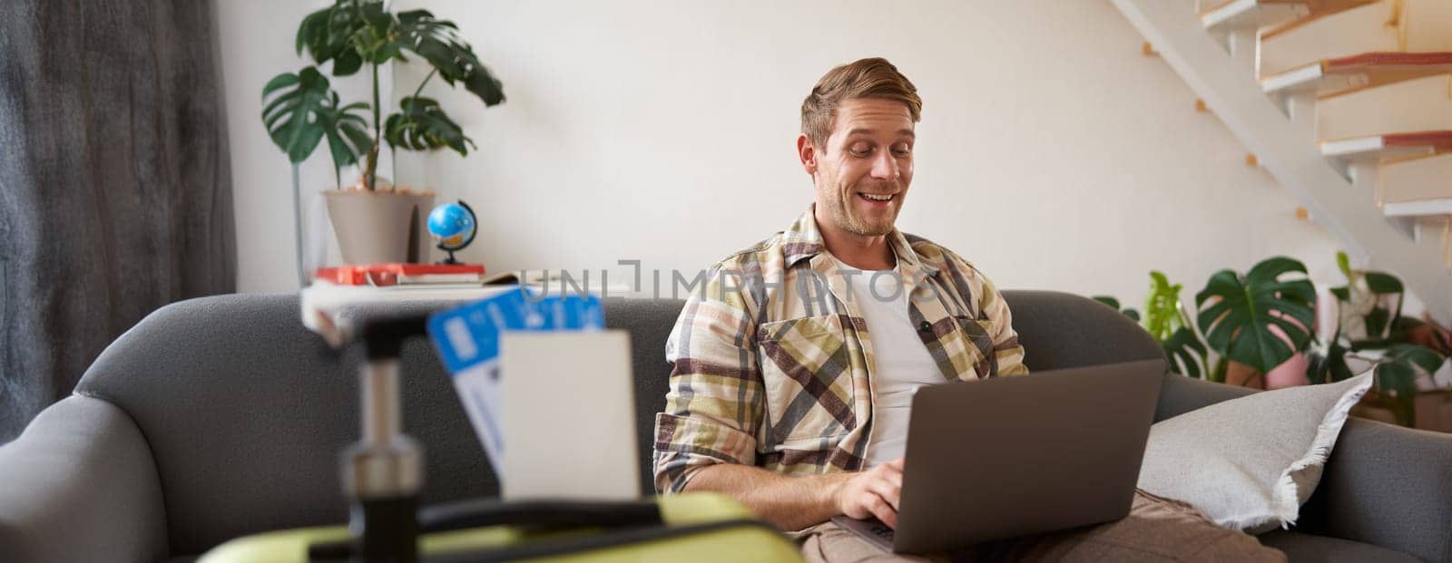 Portrait of happy young man booking holiday for two, has plane tickets, looks at laptop, sits in living room with computer. Travelling and tourism concept