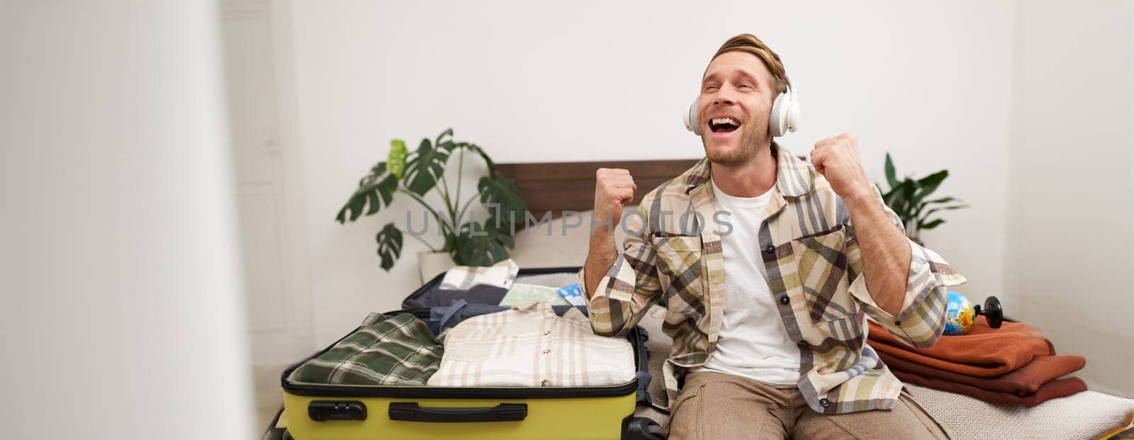 Excited young man in headphones, gets ready for vacation, goes on holiday, packs things in suitcase, listens to music in earphones, sits near his luggage. Tourism and lifestyle concept