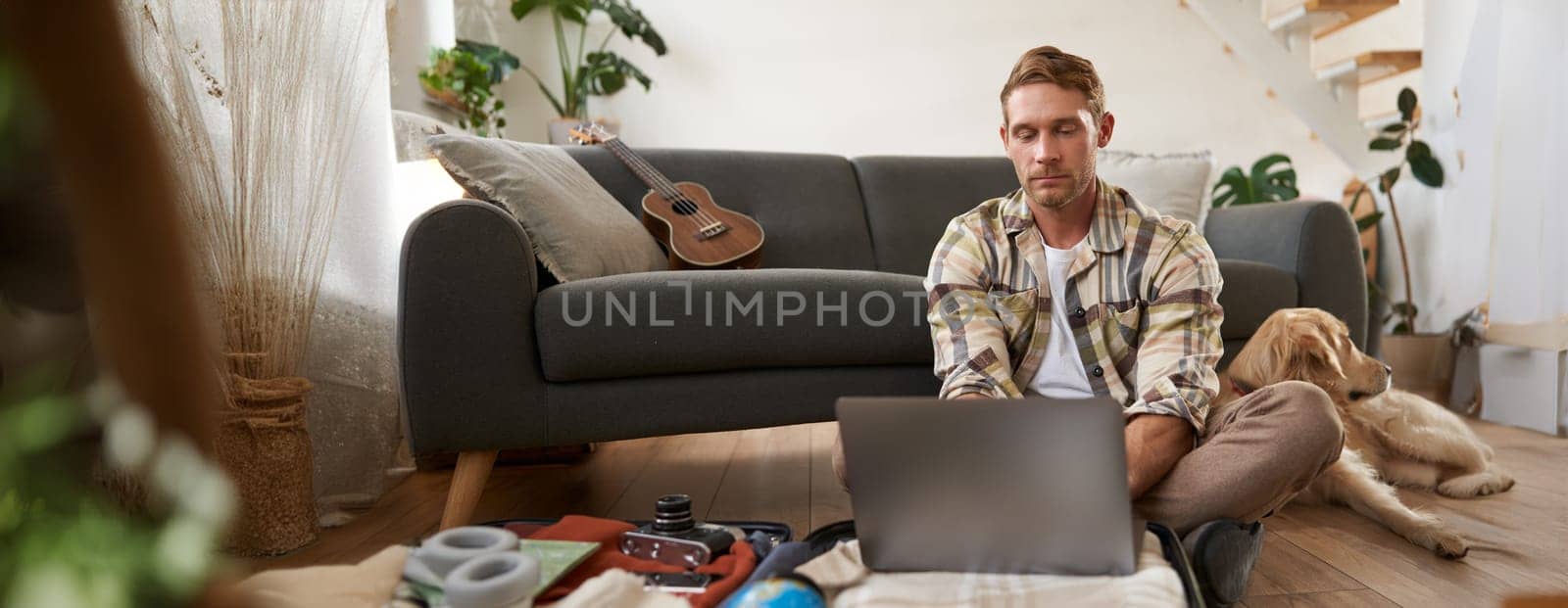 Man with laptop on suitcase, sits with dog on floor, looks online, browsing websites, packs clothes for vacation, goes on holiday, plans trip abroad.