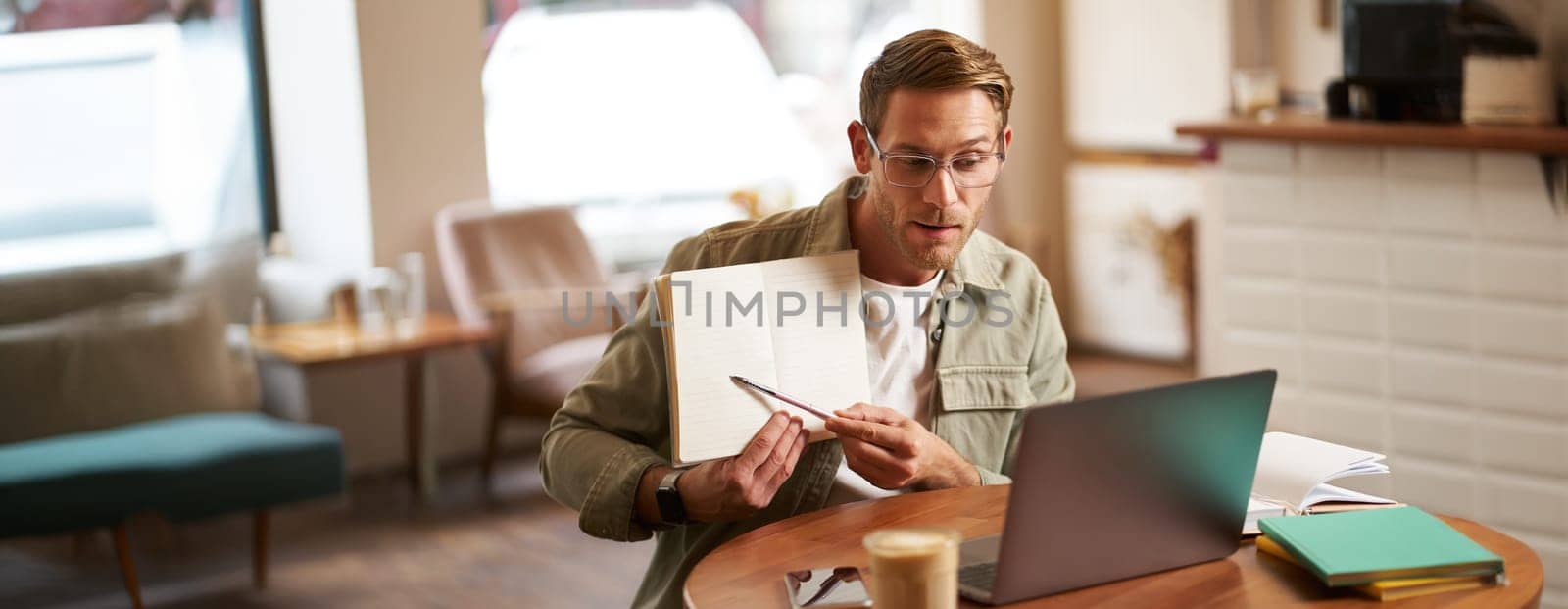 Portrait of young handsome man in glasses, private tutor teaching student online, pointing at his notebook, showing something, video chats via laptop application, working remotely in cafe.