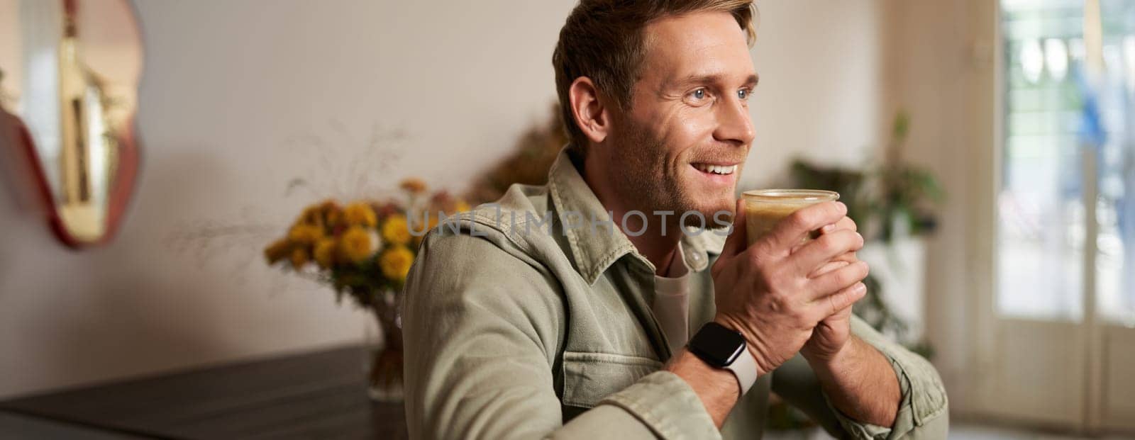 Leisure, people and lifestyle concept. Happy handsome young man in cafe, sitting on chair and smiling, holding glass of coffee, looking aside, relaxing with cup of cappuccino.