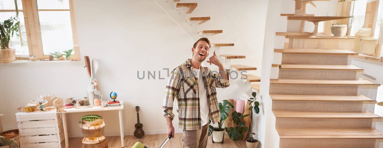 Vertical portrait of happy young man going on holiday, shows mobile phone call hand gesture, walking with suitcase, leaving rented apartment. Travelling and tourism concept