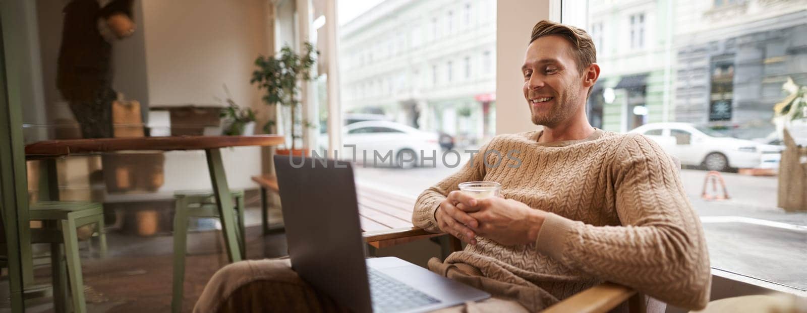 Portrait of handsome young man sits in cafe, drinks coffee and watches video on laptop, looking at screen with happy smile, relaxing in co-working space.