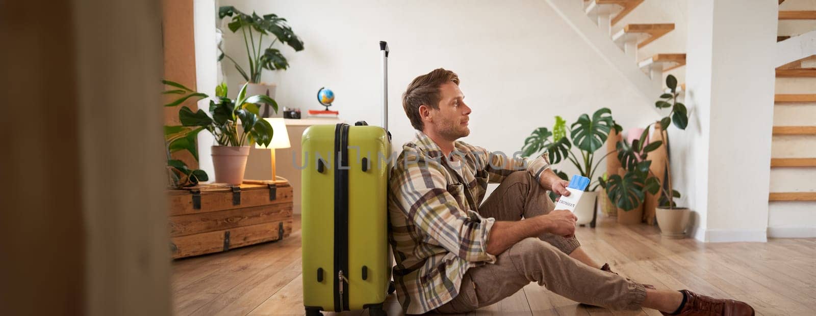 Portrait of smiling handsome tourist, man holding passport and flight tickets, sitting with suitcase. Tourism and travelling concept