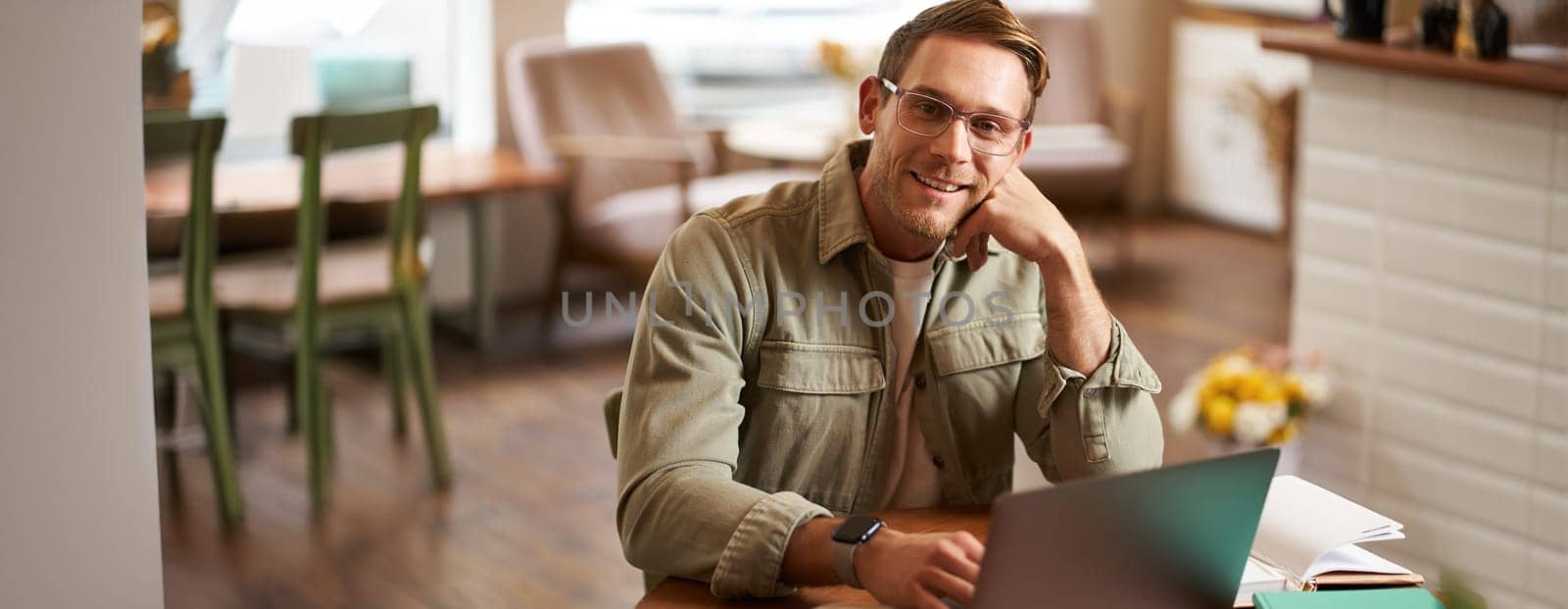 Remote work and freelance concept. Handsome man in glasses sits with notebook and laptop, studying online, working remotely from coffee shop, sitting in cafe with computer.