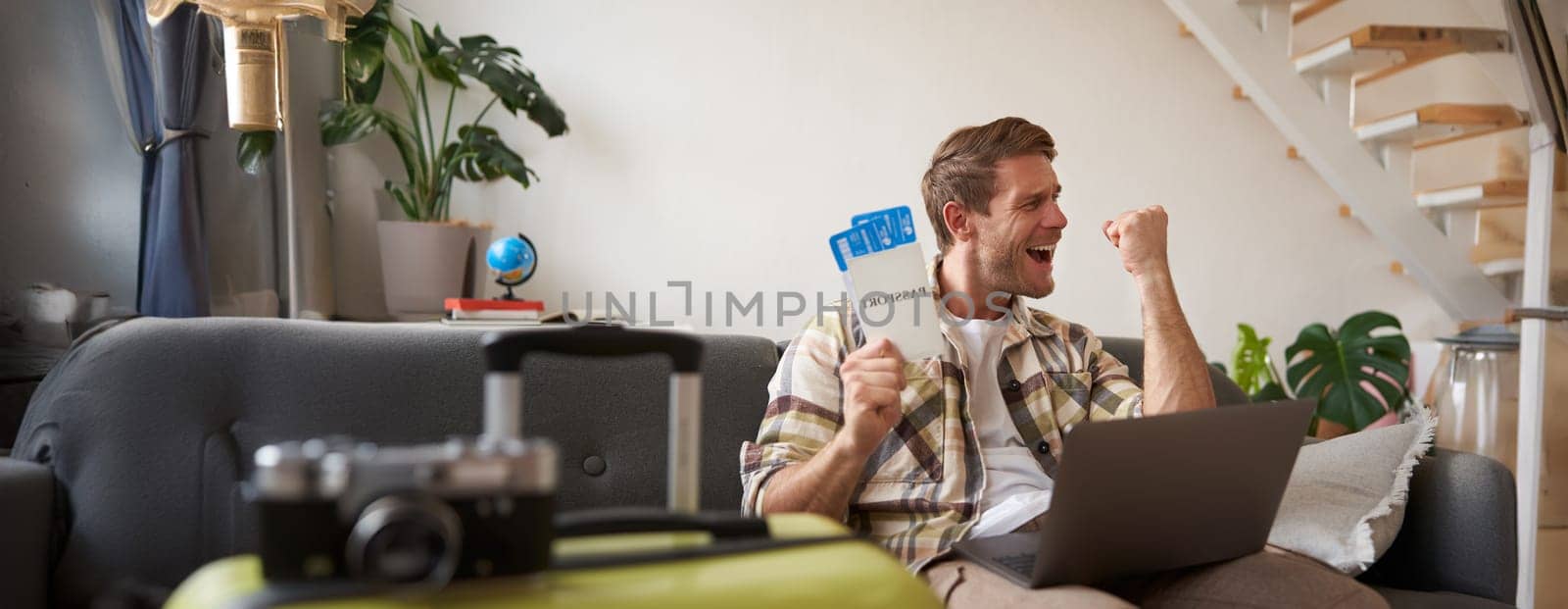 Upbeat man, tourist with two tickets, makes a fist pump, has laptop opened, goes on vacation, excited about holiday. Copy space