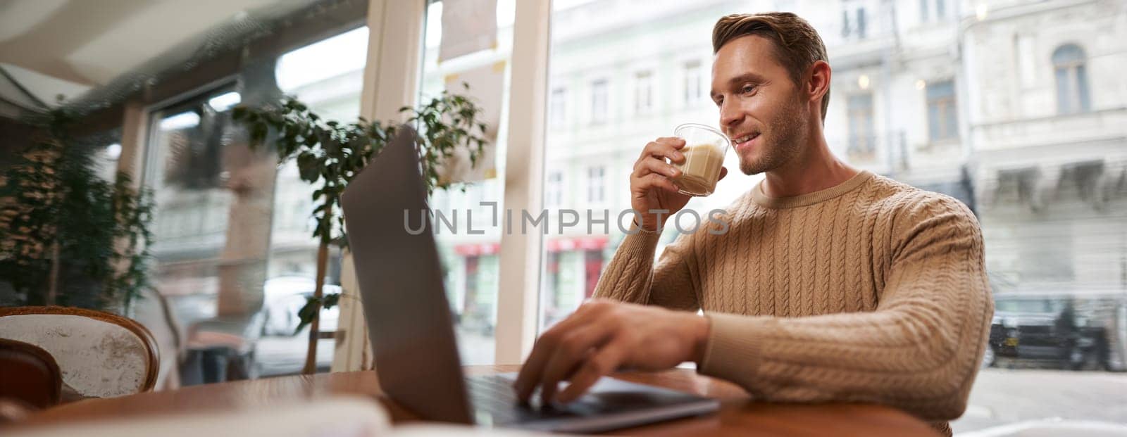 Portrait of young male businessman in cafe, drinking coffee and using his laptop, working from co-working space, sitting near the window and looking at computer screen.