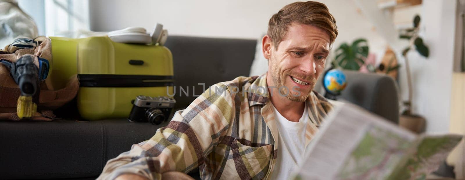 Smiling handsome man with map in hands, packed suitcase, goes on holiday, prepared to go on vacation.
