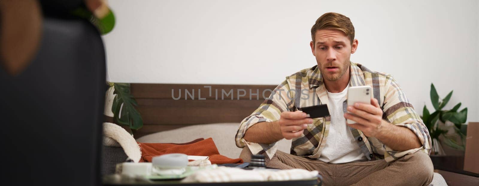 Portrait of young man, tourist paying online, using credit card and mobile phone app, sitting on bed with suitcase, ordering in internet, buying holiday tickets.