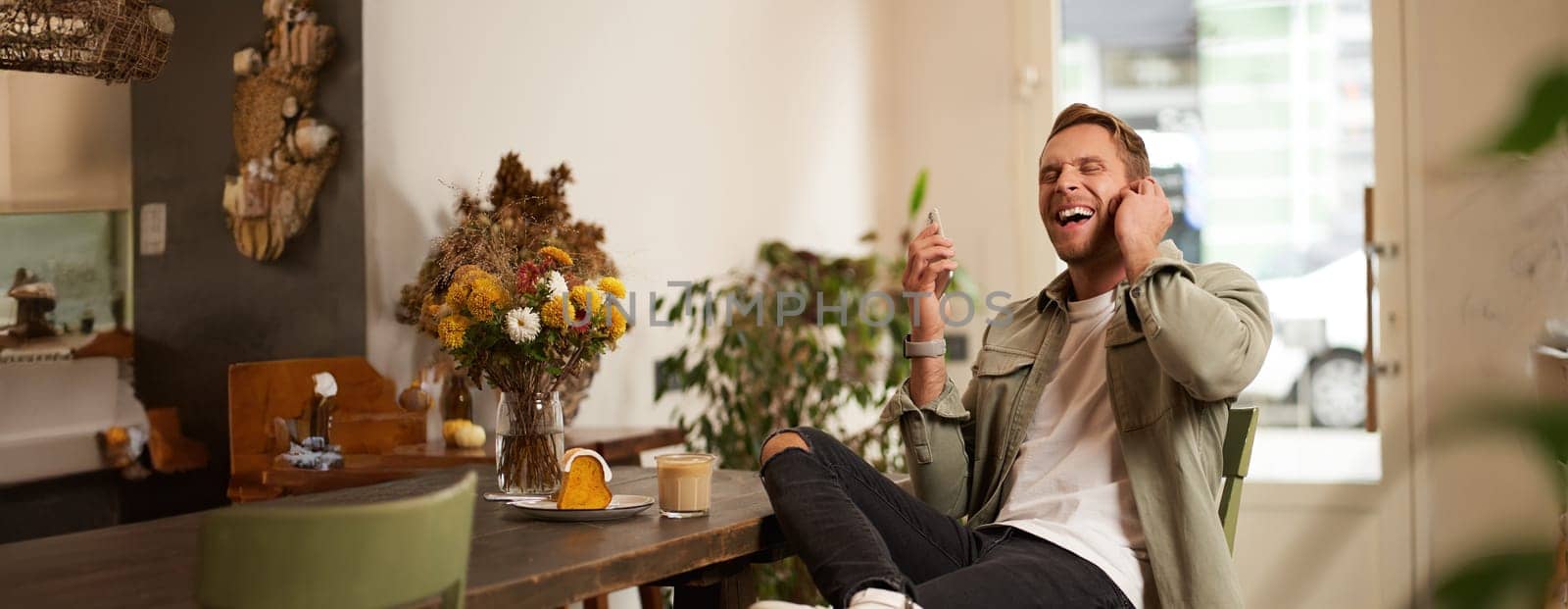 Lifestyle portrait of handsome young man, sitting in cafe, talking to a friend on mobile phone with wireless earphones, laughing and smiling, listening to funny podcast.