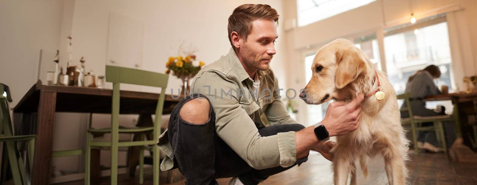 Portrait of happy man spending time with his dog in pet-friendly cafe, playing and petting golden retriever, having fun, giving him a treat.