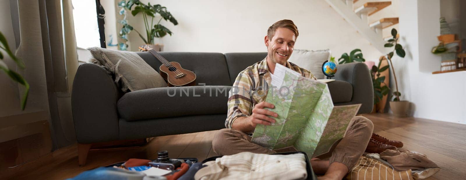 Portrait of handsome young man, traveller with suitcase, looking at paper map, choosing where to go on vacation, going on holiday, choosing route.