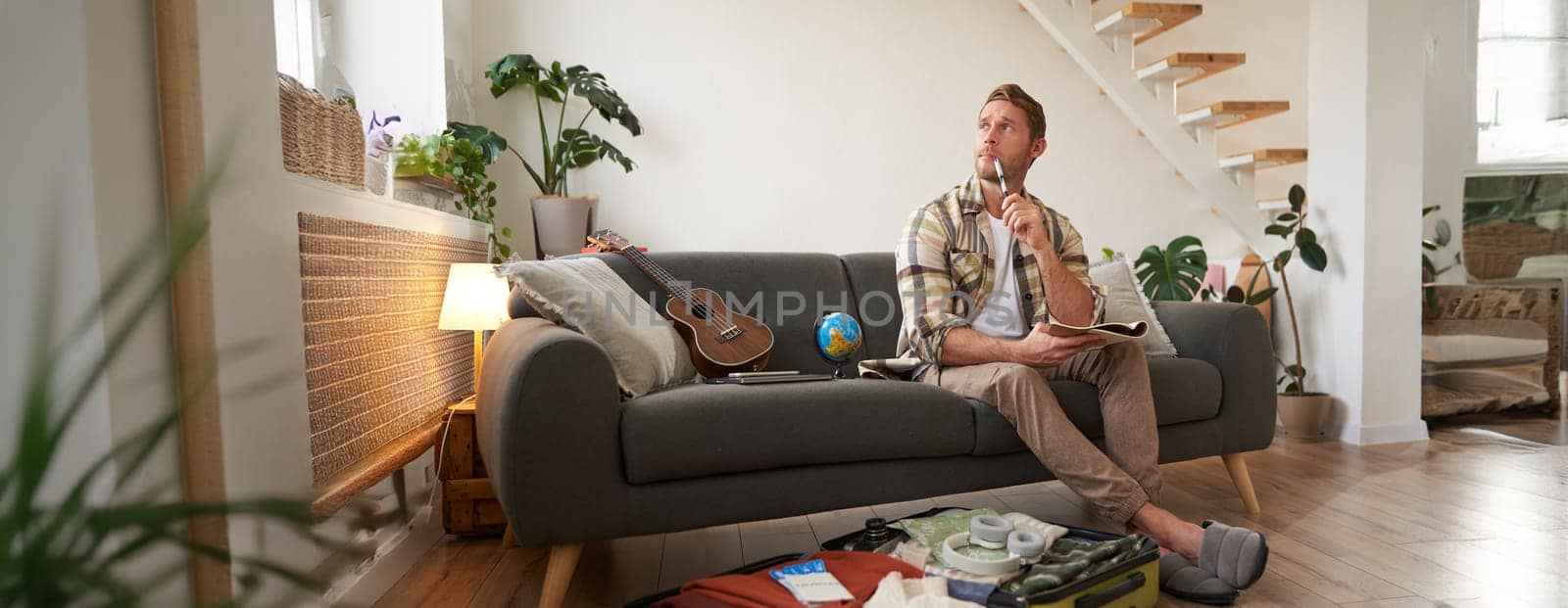 Portrait of man thinking what to take with him on vacation, holding notebook, writing list of items with thoughtful face, preparing to go on holiday, sitting near suitcase with clothes.