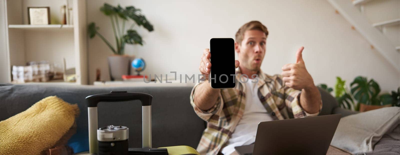 Portrait of handsome young man, showing smartphone screen, recommending mobile app for travelling or booking online, sitting with laptop and suitcase. Copy space