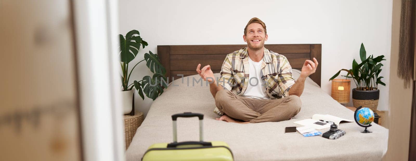 Close up portrait of meditating tourist, man sits with suitcase in bedroom, practice yoga, relaxing, finding nirvana. Tourism and travelling concept