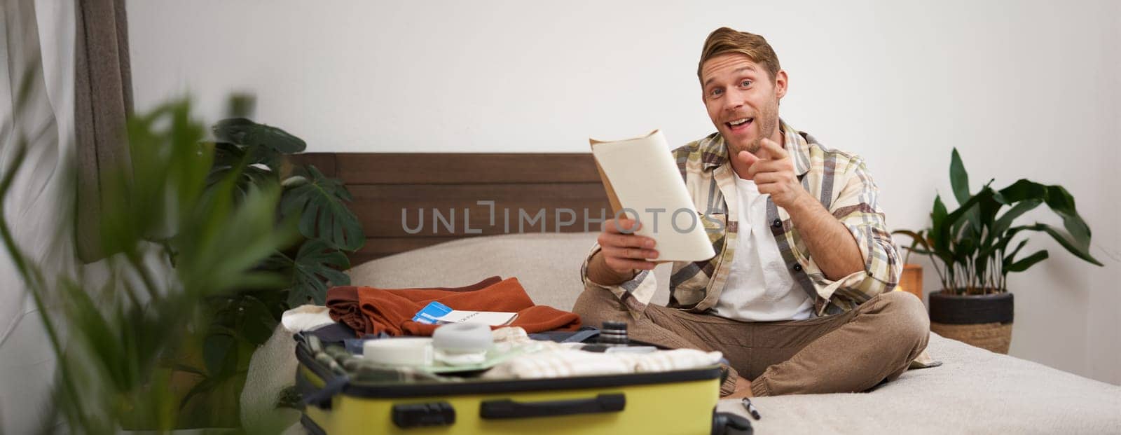 Portrait of happy, charismatic young man, excited about holiday, holding notebook, packing suitcase with clothes for vacation, pointing finger at camera and smiling.
