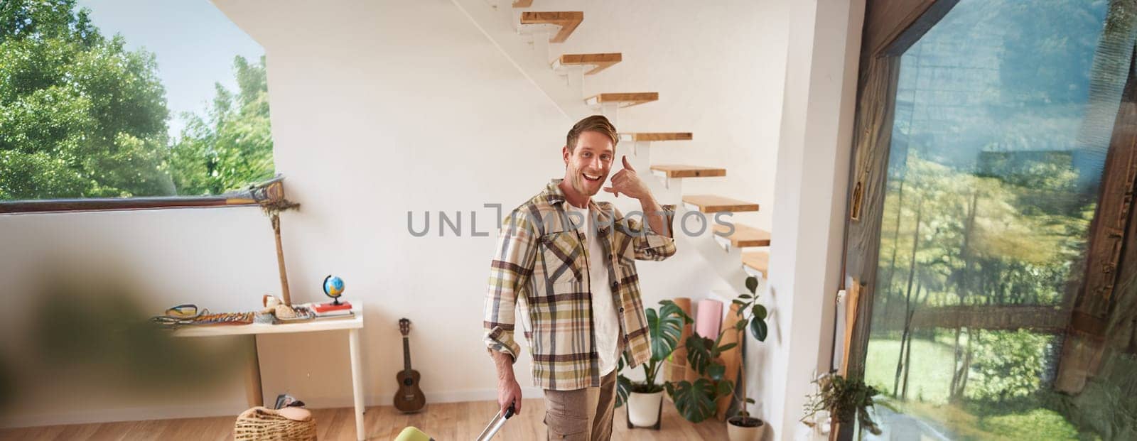 Full length shot of happy traveller, tourist goes on vacation, walks with suitcase, shows smartphone call gesture, leaves the apartment. Copy space
