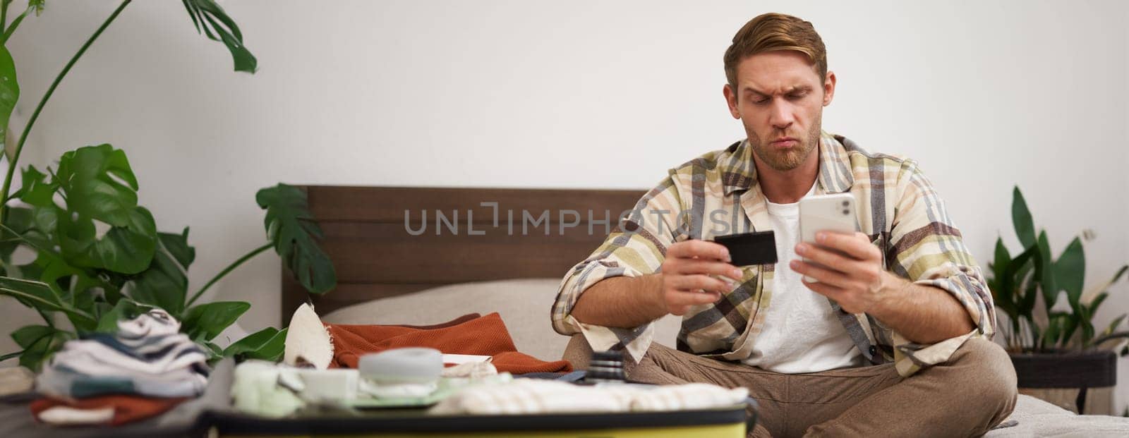 Portrait of man with thoughtful face, sitting on bed with suitcase, paying online with credit card and smartphone app. Copy space