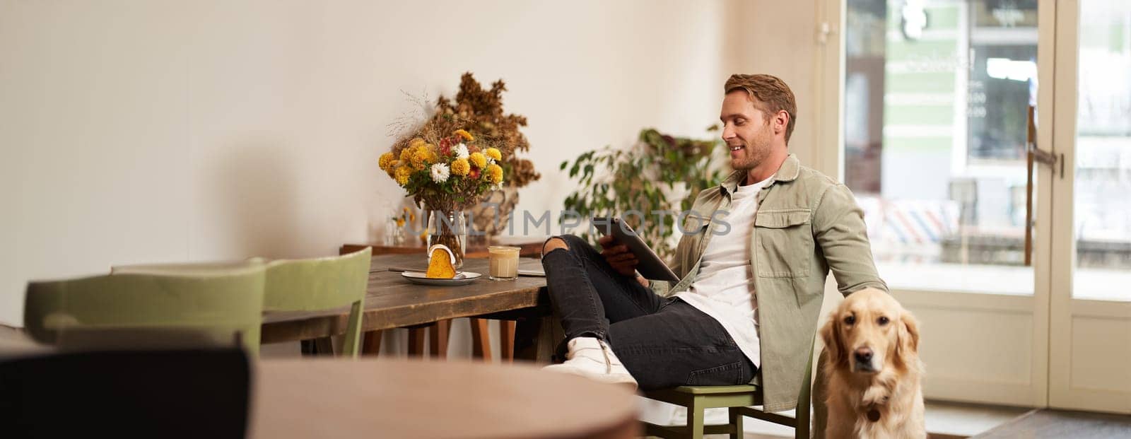 Portrait of handsome young man sits in cafe with his dog, petting golden retriever, reading news on tablet, relaxing with cup of coffee.