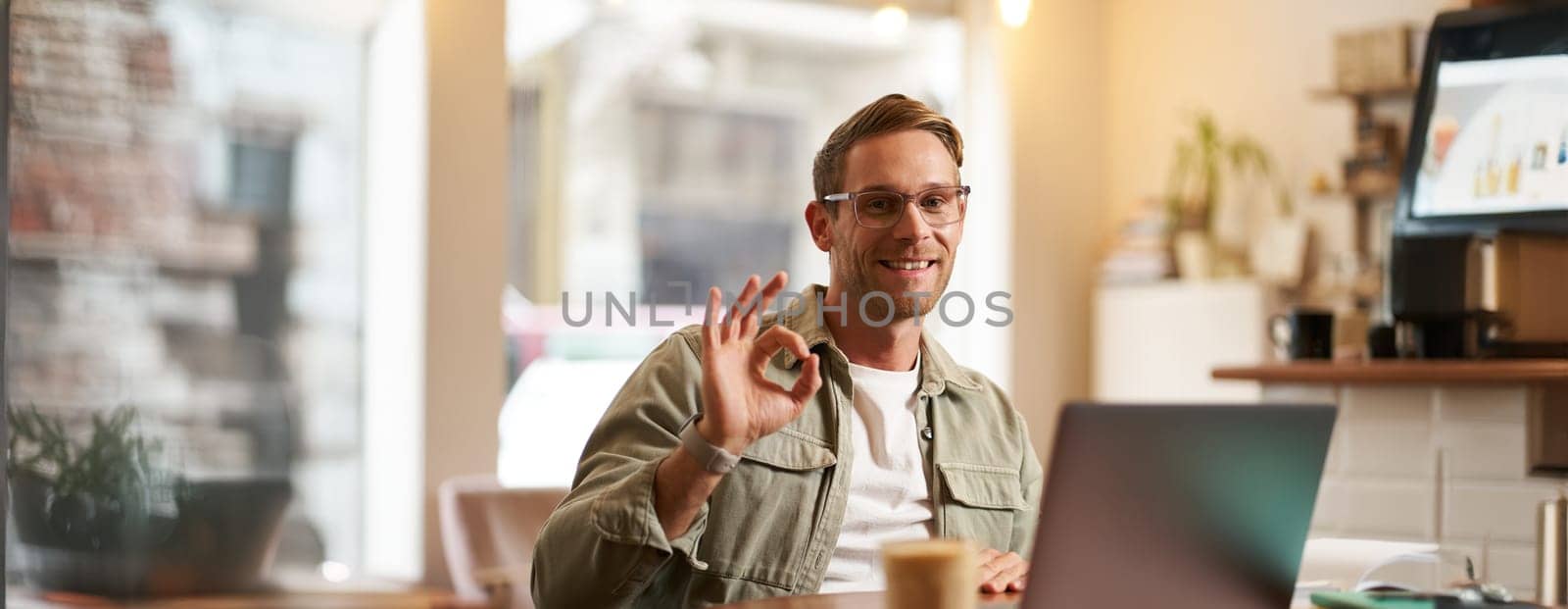 Portrait of handsome businessman in glasses, showing okay hand sign, sitting with laptop in cafe, working remotely, recommends co-working space.
