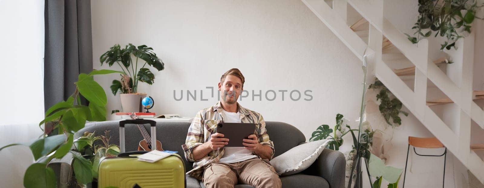 Vertical shot of young man going abroad, packing suitcase for a vacation, holding digital tablet, booking hotel online. Tourism and travel agency concept
