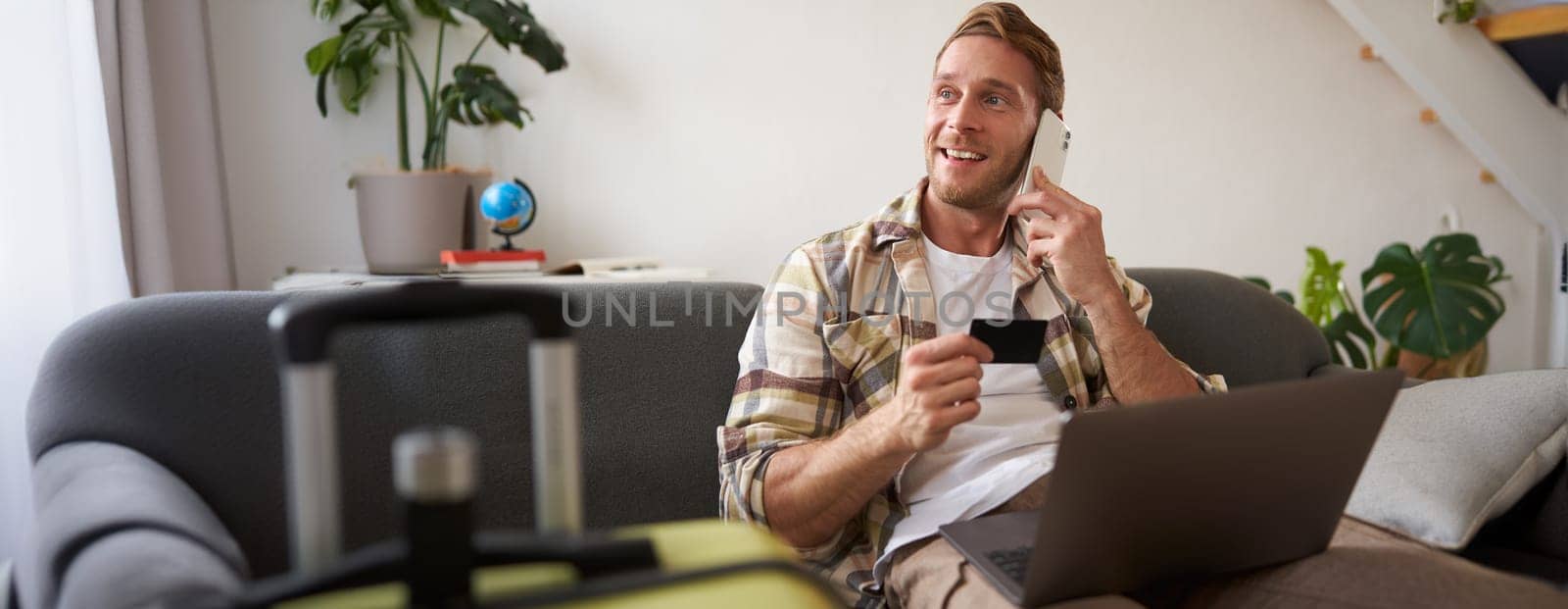 Portrait of young man with credit card, calling to book tickets, using laptop, preparing for travelling abroad, going on holiday. Tourism and finance concept
