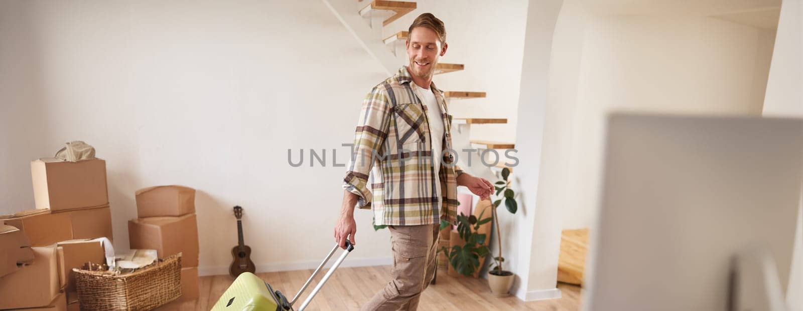 Vertical portrait of happy young man with suitcase, walking indoors, leaving a flat with luggage, going on business trip or vacation. Copy space