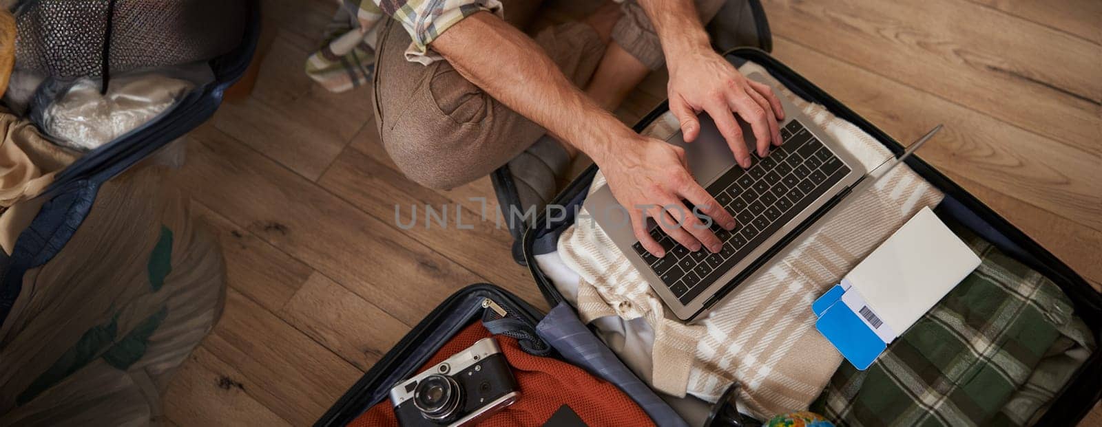 Close up shot of male hands, tourist using laptop, sitting on floor with opened suitcase, packed luggage for holiday, has two flight tickets, camera and clothes.