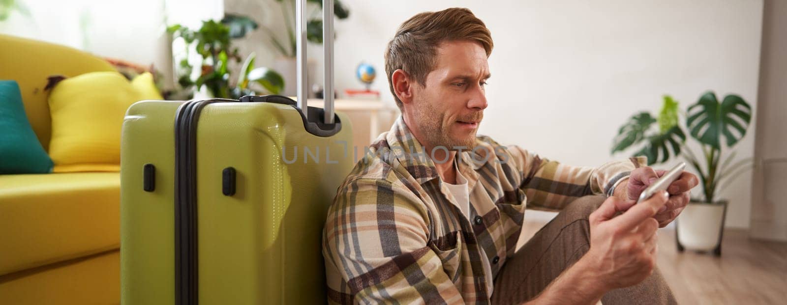 Portrait of man with suitcase, sits on floor, plays on his mobile phone, looks serious. Tourism and people concept