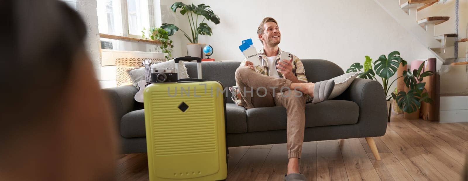 Handsome young man sits on sofa with suitcase, holds smartphone, books hostel or holiday vacation, chats on mobile phone. Tourism and travelling concept